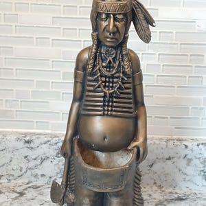 May include: Bronze-colored Native American figure with a headdress, holding a small axe and a pouch. The figure has detailed facial features and is wearing a vest and beaded necklaces. The figure is standing on a countertop.