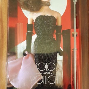 May include: A vintage Barbie doll in a clear display case. The doll wears a black sequined dress, long black gloves, and a choker. The box reads "ORIGINAL 1960 FASHION & DOLL! Special Edition Reproduction" and features the words "Solo in the Spotlight" and "Barbie".