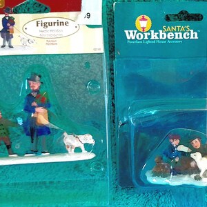 May include: Two Christmas figurines in plastic packaging. One features a man, a child, and a dog. The other shows two children with a dog. The packaging includes the text "Figurine" and "Santa's Workbench".
