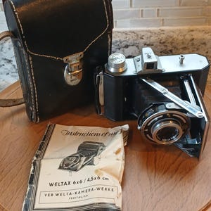May include: A vintage black and silver camera with a leather case and instruction manual. The camera is a WELTAX 6x6 / 4.5x6 cm model. The instruction manual is white with black text and a diagram of the camera.