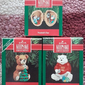 May include: Three Hallmark Keepsake Christmas ornaments in their original boxes. The boxes are red and green. One ornament is a bear holding a Christmas tree, another is a polar bear wearing a red sweater that says "DAD 1991", and the third is a nutshell with two figures inside.