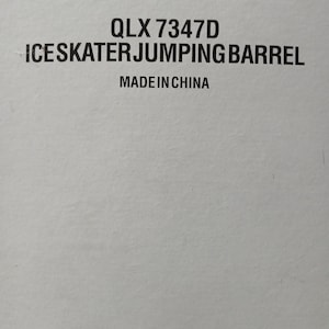 May include: A close-up of a white surface with black text. The text reads "QLX 7347D ICESKATER JUMPING BARREL MADE IN CHINA". The text is centered and in a sans-serif font.