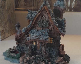 May include: A miniature wooden cabin with a stone foundation and a slate roof. The cabin has a sign that reads "The Bears Den". There is a small boat in front of the cabin.