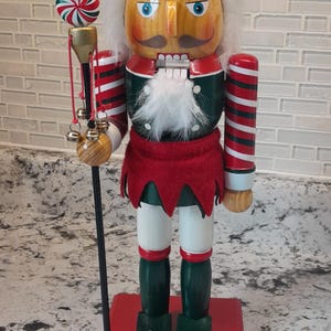 May include: A wooden Christmas nutcracker in the shape of an elf, with a green hat, red and white striped arms, and a red base. The nutcracker holds a staff with a candy cane ornament. The base has the number "2005".