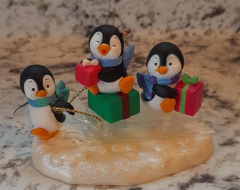 May include: A decorative figurine featuring three cartoon penguins with black and white bodies, orange feet, and blue scarves. Each penguin holds a wrapped gift. The figurine sits on a light-colored base with glitter.