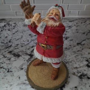 May include: A figurine of Santa Claus in a red suit with white faux fur trim, brown boots, and a belt. Santa is holding a brown glove and smiling. The figurine is standing on a round, textured base.