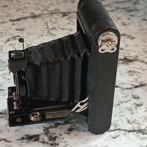 May include: A vintage black folding camera with a bellows design. The camera has a leather-like texture and a silver clasp. The text "Eastman Kodak Stores INCORPORATED PORTLAND" is visible on the camera.
