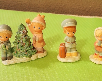 May include: A collection of small figurines depicting children in winter attire and a Christmas tree. The figurines are in pastel colors and have a vintage, collectible aesthetic. One figure holds a star, another a suitcase.