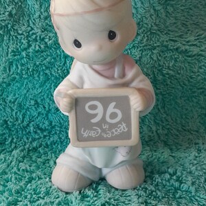 May include: A figurine of a child holding a sign that reads "96 in peace on earth." The figurine is wearing a white outfit and has a silver halo. The background is a teal, textured surface.