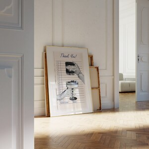 May include: A framed print with a gold border leans against a white wall. The print features a vintage-style receipt with illustrations of cocktails and the text "Thank You!". The image is in black and white with blue lines.
