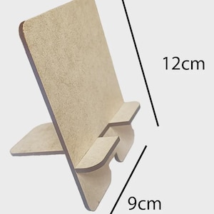 Puede incluir: Soporte de teléfono de madera marrón claro, de 12 cm de alto y 9 cm de ancho. El soporte tiene una parte trasera rectangular y una pequeña repisa para sujetar un teléfono. Diseñado para sostener un teléfono en posición vertical.