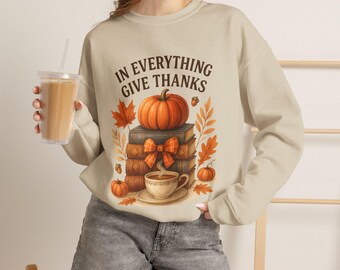 May include: A beige crewneck sweatshirt featuring a graphic of a stack of books topped with an orange pumpkin, a plaid bow, and a steaming cup of coffee, with the text IN EVERYTHING GIVE THANKS and autumn leaves surrounding the central design.