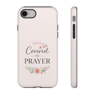 May include: A light pink phone case with the words "Covered in Prayer" in black script, with floral accents. The year "2026" is above the text. The case has a black border and a camera cutout.