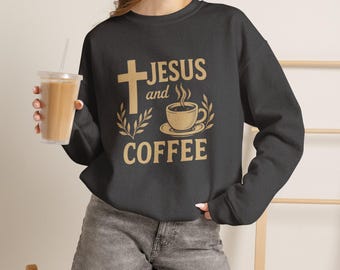 May include: A dark grey long-sleeve sweatshirt featuring a tan graphic print that reads "JESUS and COFFEE" with an illustration of a cross, a steaming cup of coffee, and decorative leaf accents. The sweatshirt has a crew neckline and ribbed cuffs.