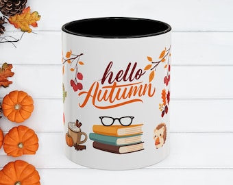 May include: White ceramic mug with a black interior, featuring the words "hello Autumn" in orange script. The mug is decorated with fall-themed illustrations, including books, glasses, a hedgehog, and pumpkins.