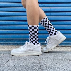 Könnte beinhalten: Weiße Sneaker mit schwarz-weißen karierten Socken. Die Sneaker haben eine weiße Sohle und ein schwarzes Swoosh-Logo.