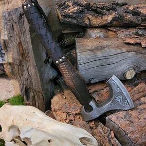 Puede incluir: Un hacha vikinga forjada a mano con un mango de madera envuelto en cuero marr&oacute;n. La cabeza del hacha presenta un dise&ntilde;o detallado con un s&iacute;mbolo r&uacute;nico y una correa de cuero. El hacha est&aacute; apoyada en una pila de madera.