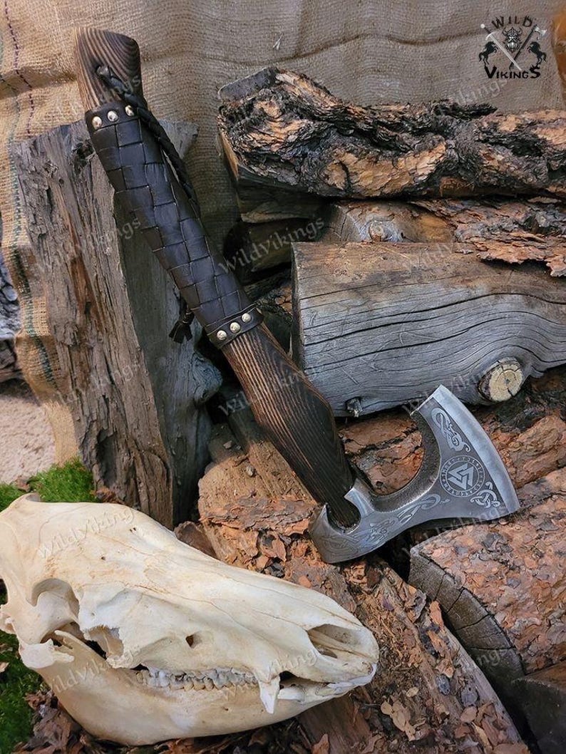 Puede incluir: Un hacha vikinga forjada a mano con un mango de madera envuelto en cuero marr&oacute;n. La cabeza del hacha presenta un dise&ntilde;o detallado con un s&iacute;mbolo r&uacute;nico y una correa de cuero. El hacha est&aacute; apoyada en una pila de madera.