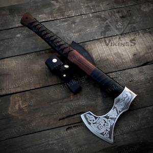 Puede incluir: Un hacha vikinga forjada a mano con un mango de madera marrón y una envoltura de cuero negro. La cabeza del hacha presenta intrincados tallados de símbolos nórdicos y un diseño de nudo celta. El hacha se muestra sobre una superficie de madera.