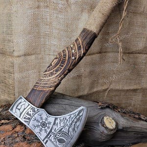 Puede incluir: Un hacha vikinga hecha a mano con un mango de madera envuelto en cuerda. La cabeza del hacha est&aacute; grabada con un dise&ntilde;o de nudos vikingos y una rosa de los vientos. El hacha est&aacute; apoyada sobre una superficie de madera.