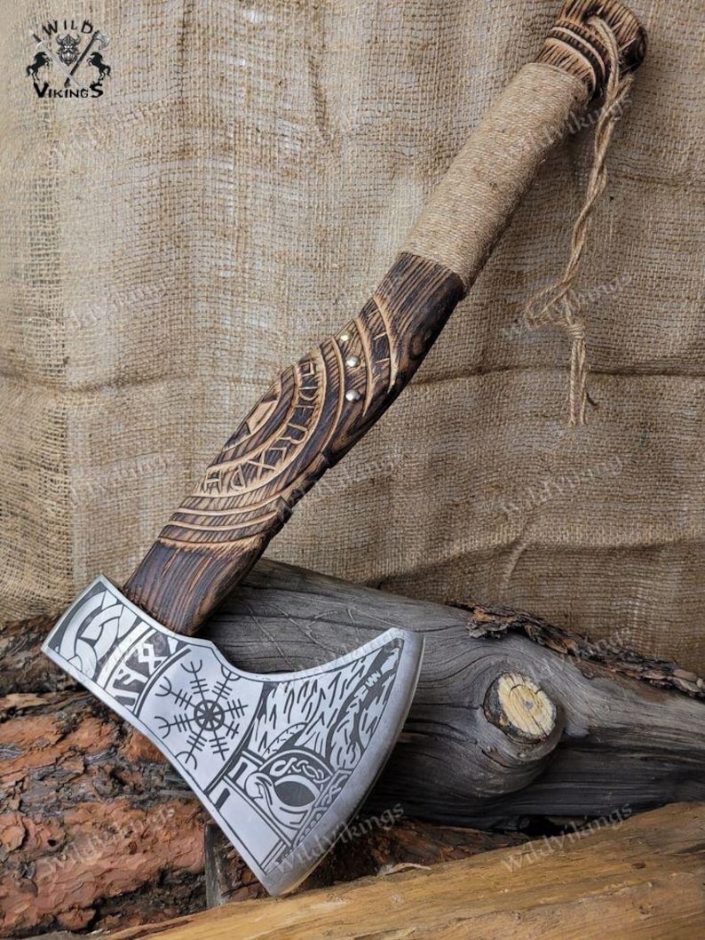 Puede incluir: Un hacha vikinga hecha a mano con un mango de madera envuelto en cuerda. La cabeza del hacha est&aacute; grabada con un dise&ntilde;o de nudos vikingos y una rosa de los vientos. El hacha est&aacute; apoyada sobre una superficie de madera.