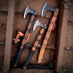 May include: Three Viking axes with leather wrappings and wooden handles. The axes have black leather grips and are decorated with runes. The axes are displayed on a rusty metal surface.
