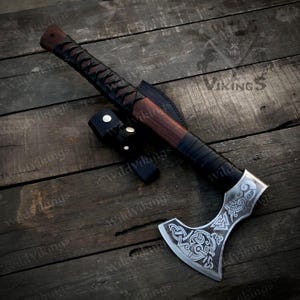 Puede incluir: Un hacha vikinga forjada a mano con un mango de madera marr&oacute;n y una envoltura de cuero negro. La cabeza del hacha presenta intrincados tallados de s&iacute;mbolos n&oacute;rdicos y un dise&ntilde;o de nudo celta. El hacha se muestra sobre una superficie de madera.