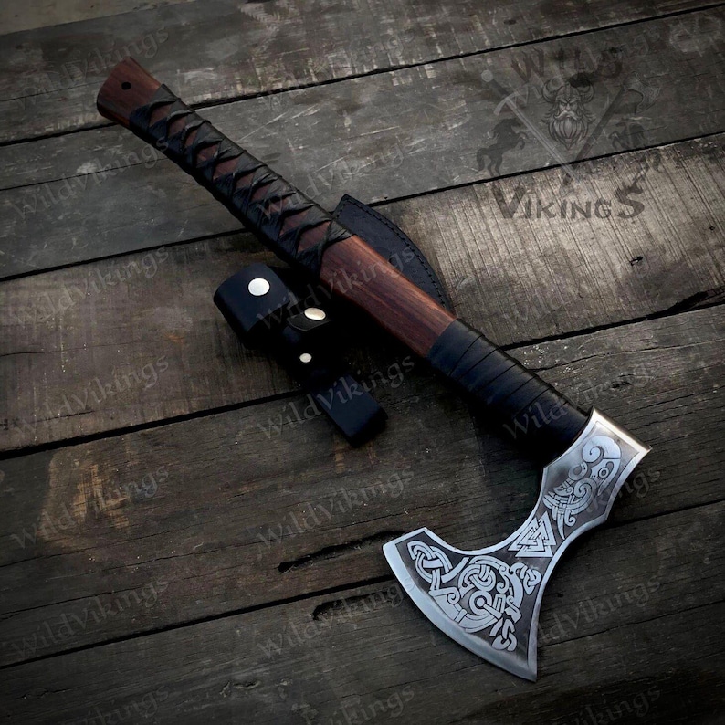 Puede incluir: Un hacha vikinga forjada a mano con un mango de madera marr&oacute;n y una envoltura de cuero negro. La cabeza del hacha presenta intrincados tallados de s&iacute;mbolos n&oacute;rdicos y un dise&ntilde;o de nudo celta. El hacha se muestra sobre una superficie de madera.