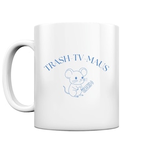Può includere: Tazza in ceramica bianca con un contorno blu di un topo che tiene un telecomando. Il testo "TRASH-TV-MAUS" è scritto in un arco blu sopra il topo.