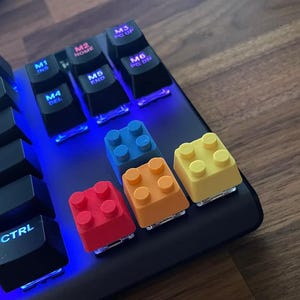 May include: A black keyboard with custom keycaps. The keycaps are designed to look like colorful toy building blocks in red, orange, blue, and yellow. Other keys are black with white lettering and blue backlighting.