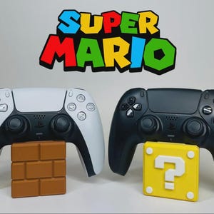 May include: Two game controllers, one white and one black, with a Super Mario logo above. The white controller rests on a brown brick block, while the black controller sits on a yellow question mark block.