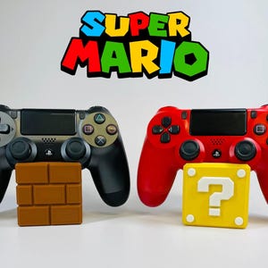 May include: Two black and red game controllers with Super Mario-themed accessories. A brown brick block and a yellow question mark block are in front of the controllers. The Super Mario logo is at the top.