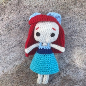 May include: A crocheted doll with red hair, a blue and white dress, and white skin.