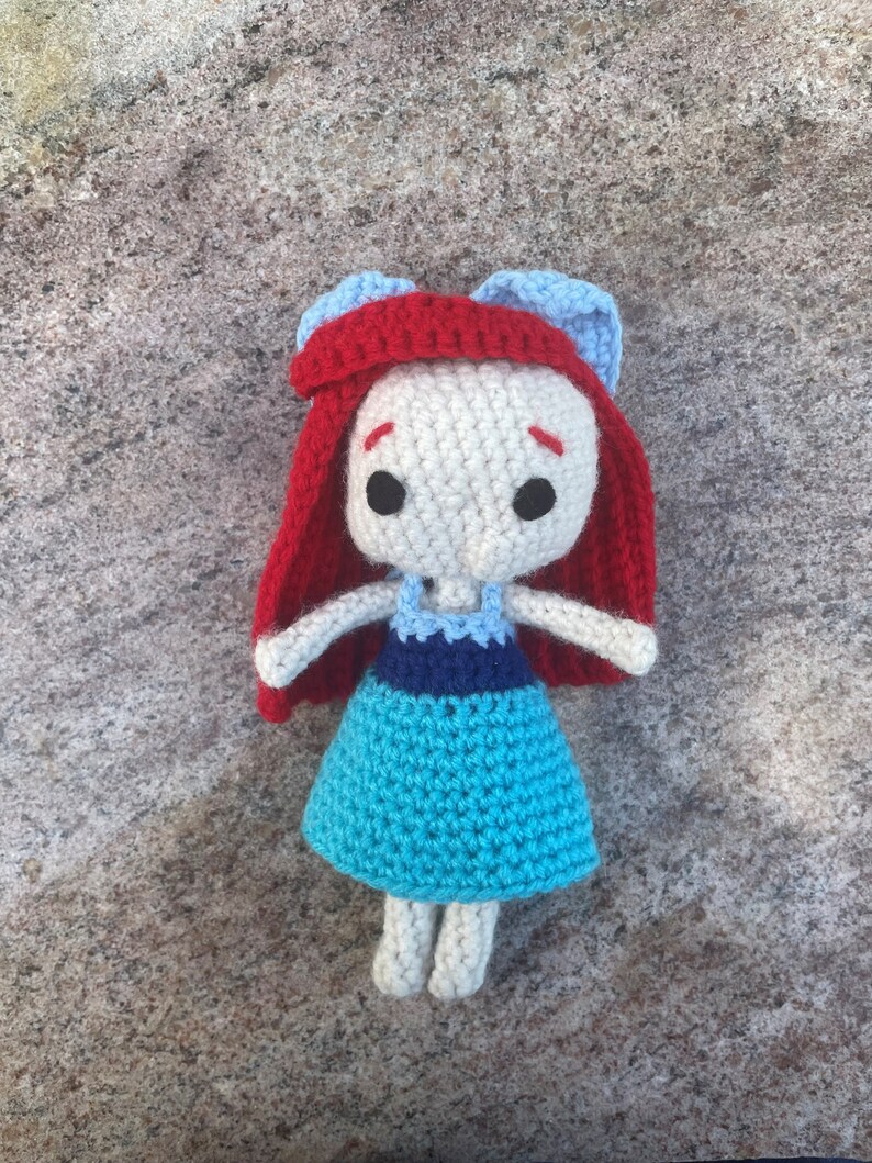 May include: A crocheted doll with red hair, a blue and white dress, and white skin.
