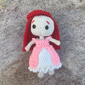 May include: A crocheted doll with red hair, a pink dress, and white crocheted details. The doll has black eyes and a white face.