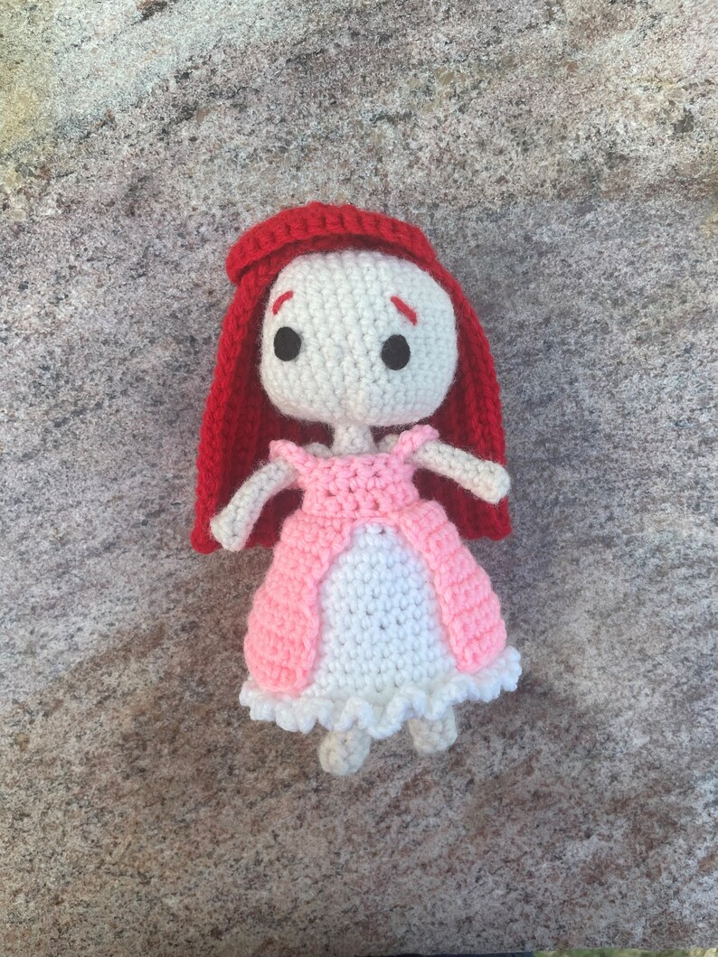 May include: A crocheted doll with red hair, a pink dress, and white crocheted details. The doll has black eyes and a white face.