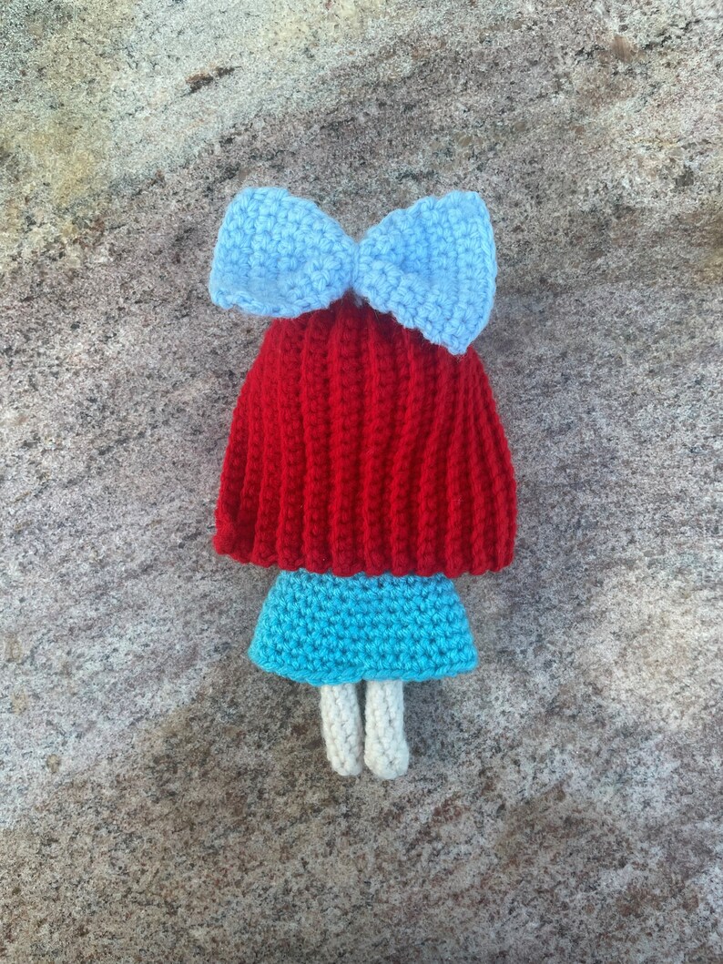 May include: A crocheted doll with red hair, a blue bow, a blue skirt, and white legs.