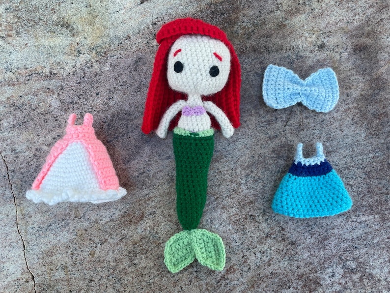 May include: A crocheted mermaid doll with red hair, a green tail, and a purple seashell bra. The doll is wearing a pink dress and a blue dress. There is also a blue bow.