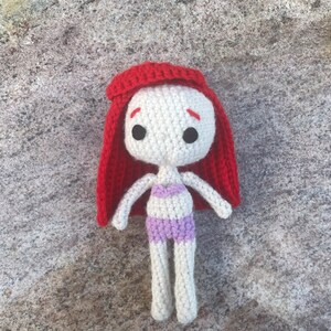May include: A crocheted doll with red hair, white skin, and a purple swimsuit. The doll is lying on a textured surface.