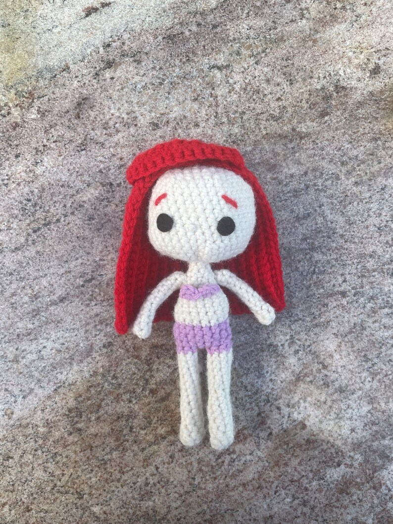 May include: A crocheted doll with red hair, white skin, and a purple swimsuit. The doll is lying on a textured surface.