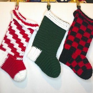 Puede incluir: Tres calcetines de Navidad de ganchillo colgados de un fondo blanco. Los calcetines son de rayas rojas y blancas, verdes y a cuadros negros y rojos.