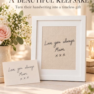 Op de afbeelding: Een witte fotolijst met een beige stoffen inzetstuk geborduurd met de zwarte tekst "Love you always Mum x x x". Een bijpassende witte kaart met dezelfde handgeschreven tekst staat voor de lijst, naast een vaas met bloemen.