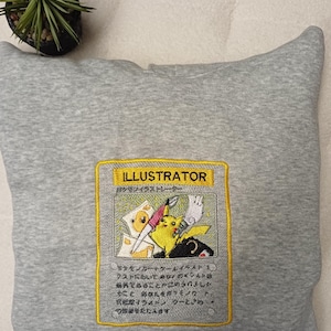 May include: A gray hoodie featuring an embroidered patch with the word "ILLUSTRATOR" in yellow. The patch depicts a cartoon character and Japanese text. A small potted plant is visible in the background.