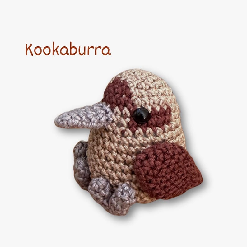 May include: A crocheted kookaburra bird in brown, beige, and grey tones. The bird features a large beak, black eyes, and small feet. The word "Kookaburra" is written in brown script above the bird.