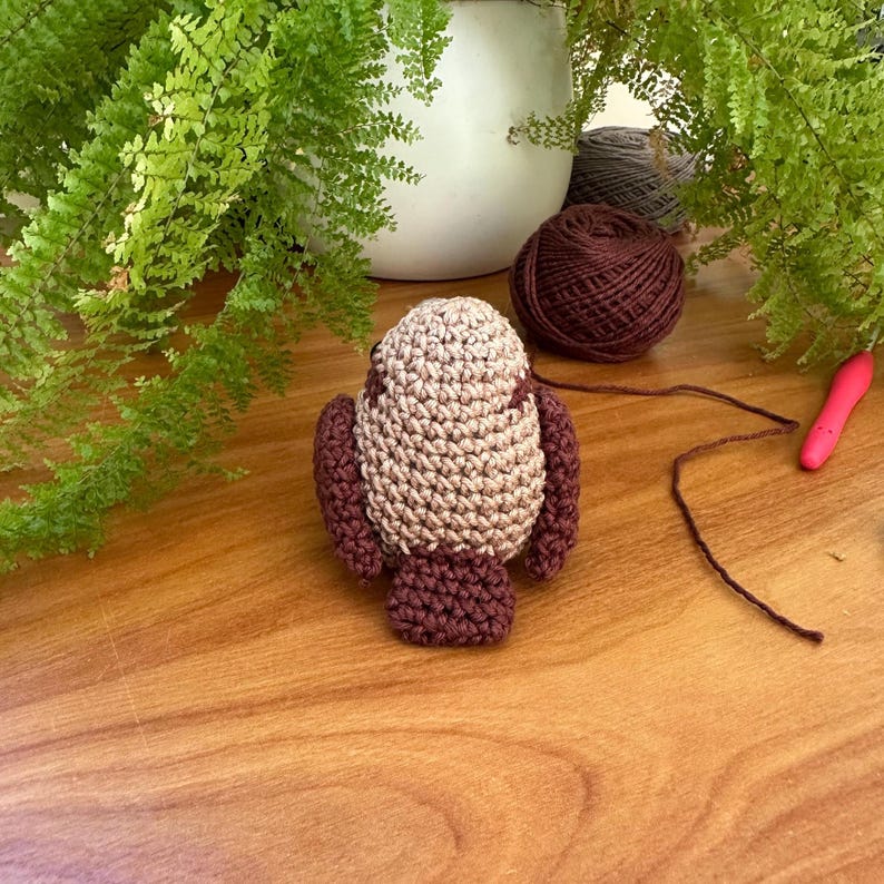 May include: A crocheted bird in tan and burgundy. The bird has a rounded body, wings, and a tail. A ball of brown yarn, a crochet hook, and greenery are in the background, suggesting a crafting theme.