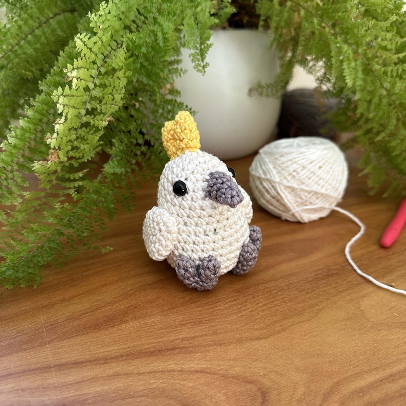 May include: A crocheted bird ornament with a white body, grey beak and feet, and a yellow crest. The bird sits on a wooden surface, with a ball of white yarn and green plants in the background. A craft project.