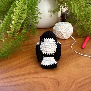 May include: A crocheted penguin in black and white, set on a wooden surface. A ball of white yarn and a pink crochet hook are also visible. Green plants and a white pot are in the background, suggesting a crafting theme.