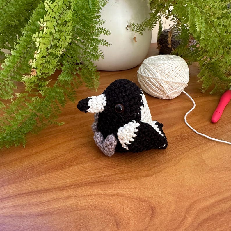 May include: A crocheted bird in black, white, and grey, with a large black beak and round eyes. The bird is on a wooden surface, with a ball of white yarn and greenery in the background.