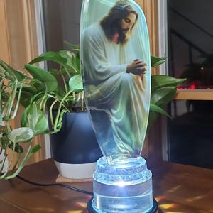 May include: A glass lamp with a clear base and a leaf-shaped shade featuring a printed image of Jesus. The lamp is illuminated with a white light, casting a warm glow on the surrounding surface.