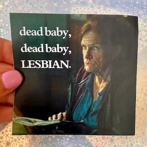 May include: A square sticker featuring a photographic portrait of an older person with a serious expression, wearing a dark jacket over a blue shirt. The text "dead baby, dead baby, LESBIAN." is printed in white, bold, sans-serif capital letters.